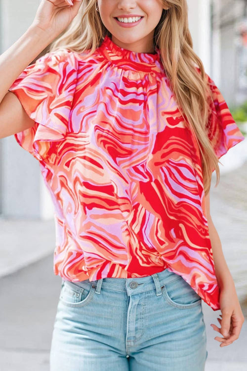 A woman wearing a red and pink abstract print ruffled short sleeve mock neck blouse with cap sleeves, paired with blue jeans.