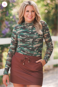 Woman wearing a camouflage long-sleeve top and brown skirt outdoors.